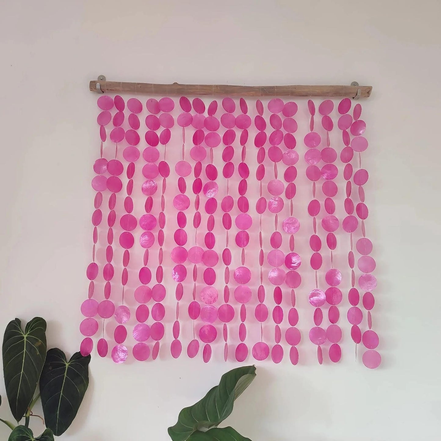 Pink capiz shell string curtain hanging on a wooden rod against a white wall with green leaves at the bottom.