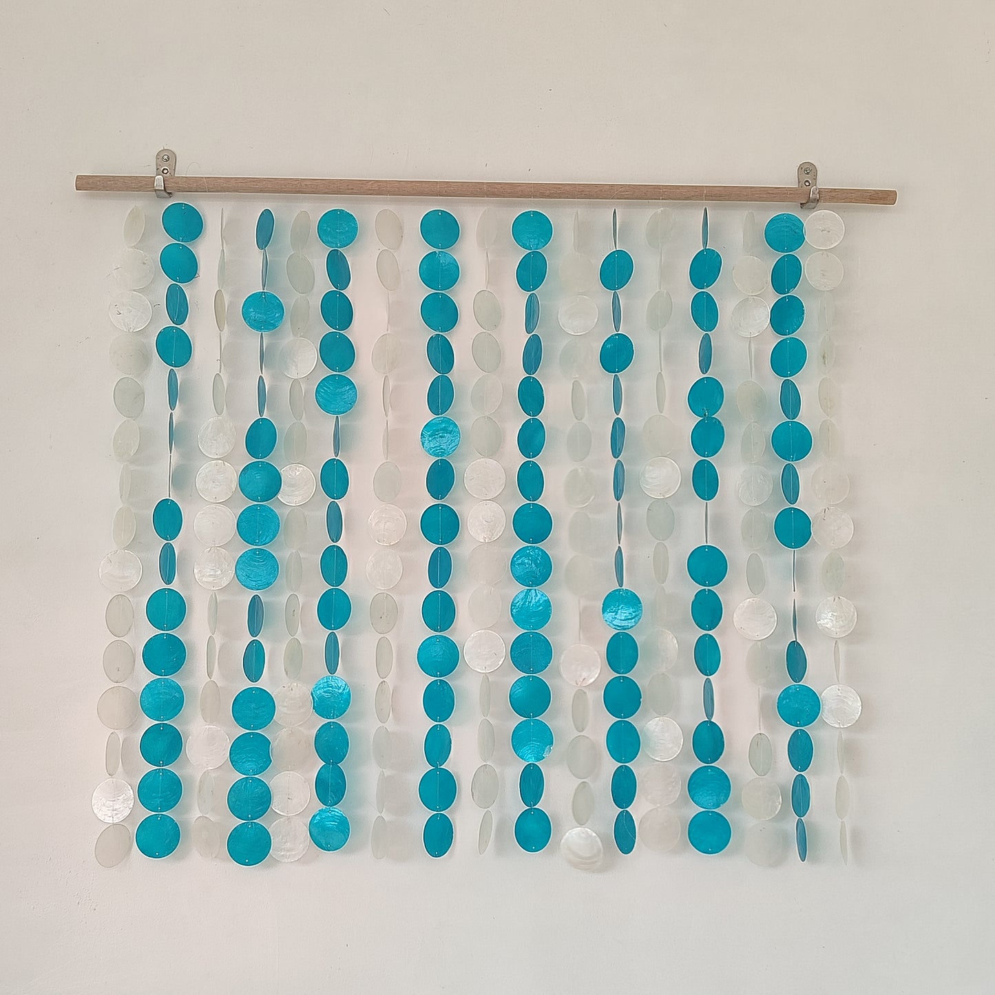 Capiz wall hanging with blue and white color hanging from a rod against a plain wall.