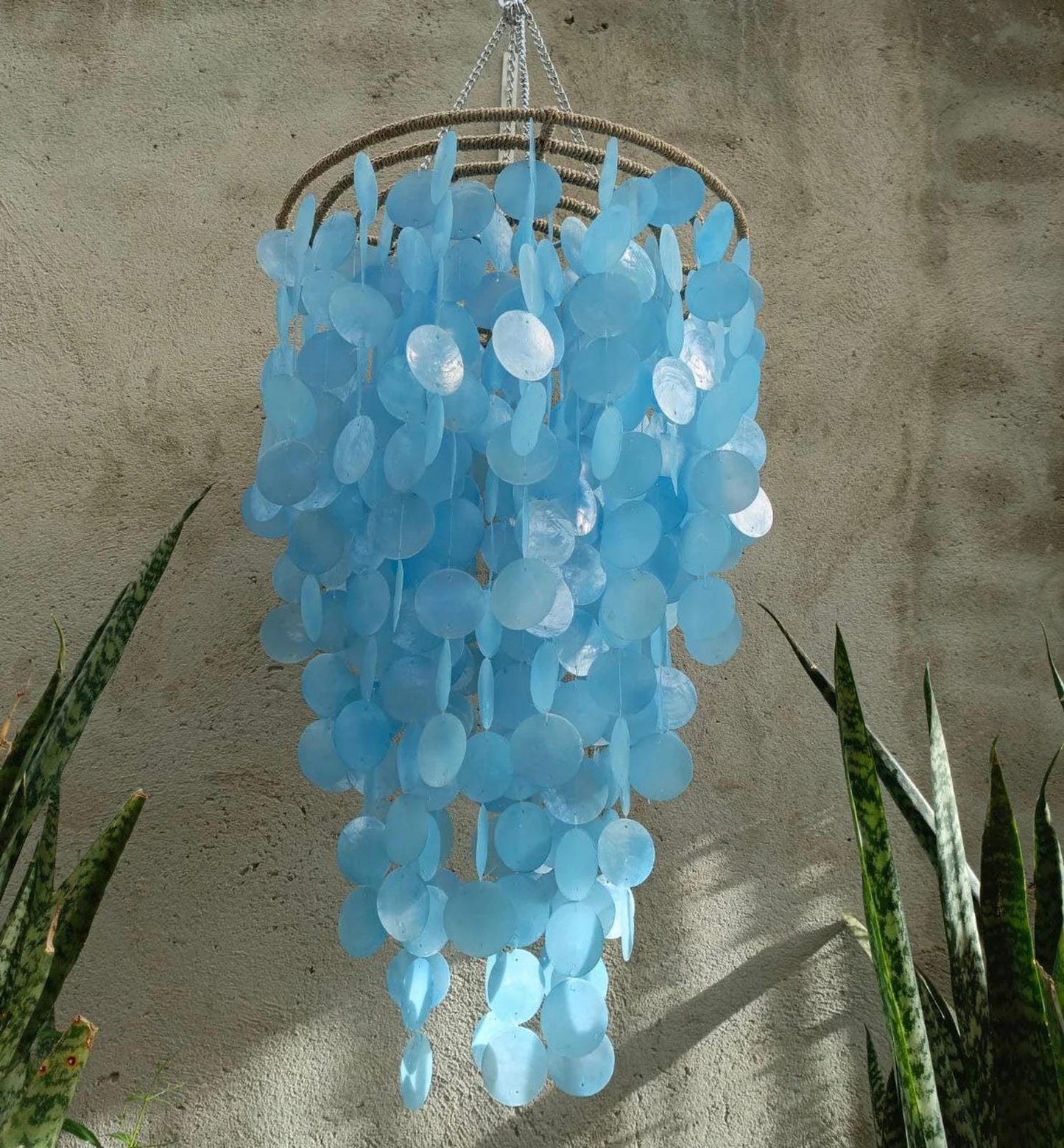 Blue capiz chandelier hanging against a textured wall with plants at the base.