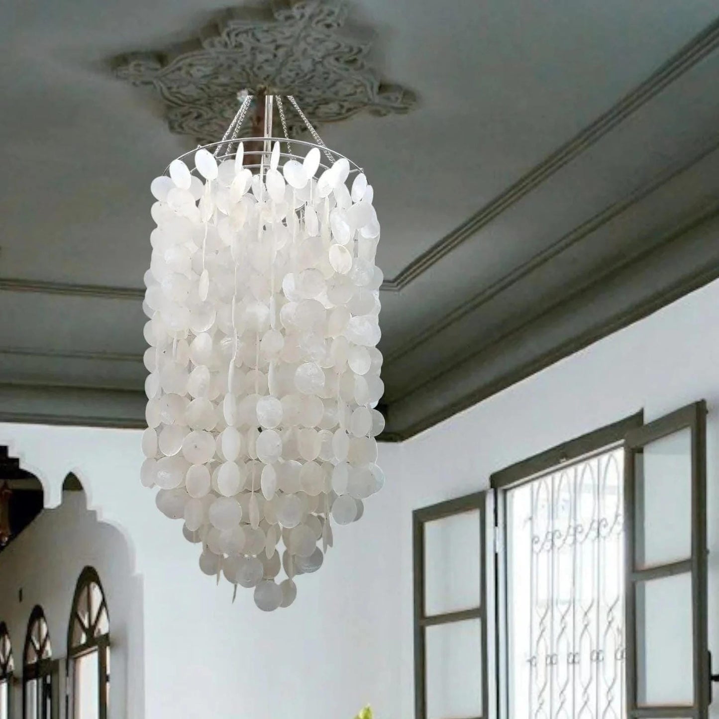 White capiz shell chandelier hanging in a room with decorative ceiling and window.
