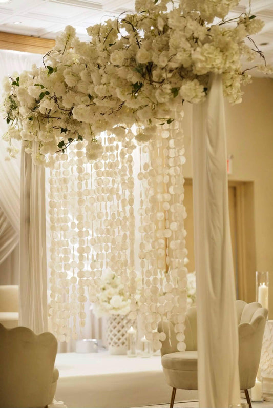 A large white floral canopy with hanging capiz shell and draped white fabric in an indoor event space.