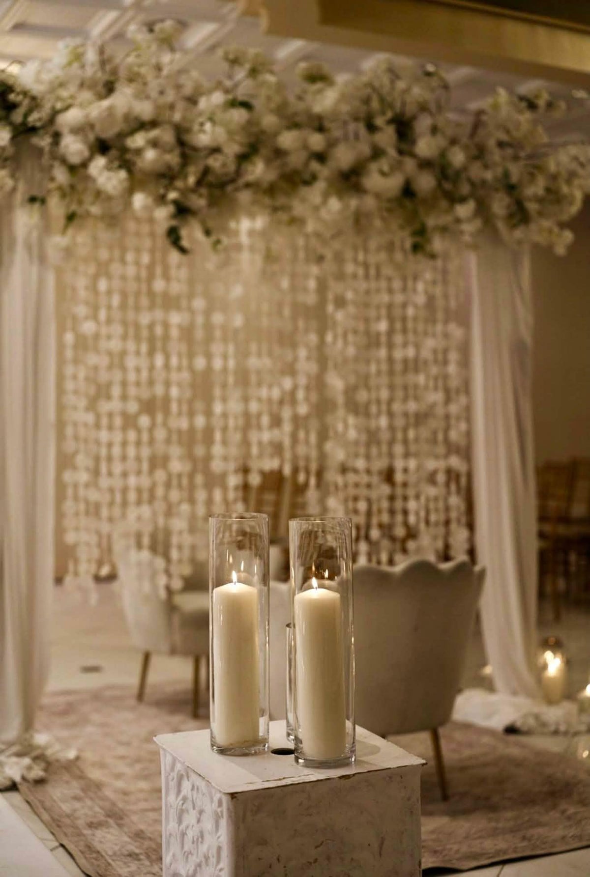 A wedding-style event setup with a floral arch, draped white fabric, hanging capiz shell, and two tall glass candle holders on a pedestal in the foreground.