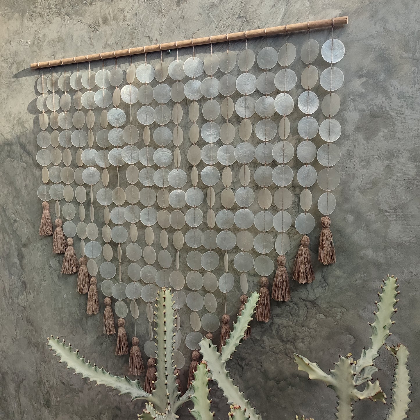 Decorative wall hanging made from capiz shell and tassels on a textured wall.