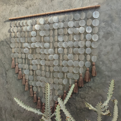 Decorative wall hanging made from capiz shell and tassels on a textured wall.