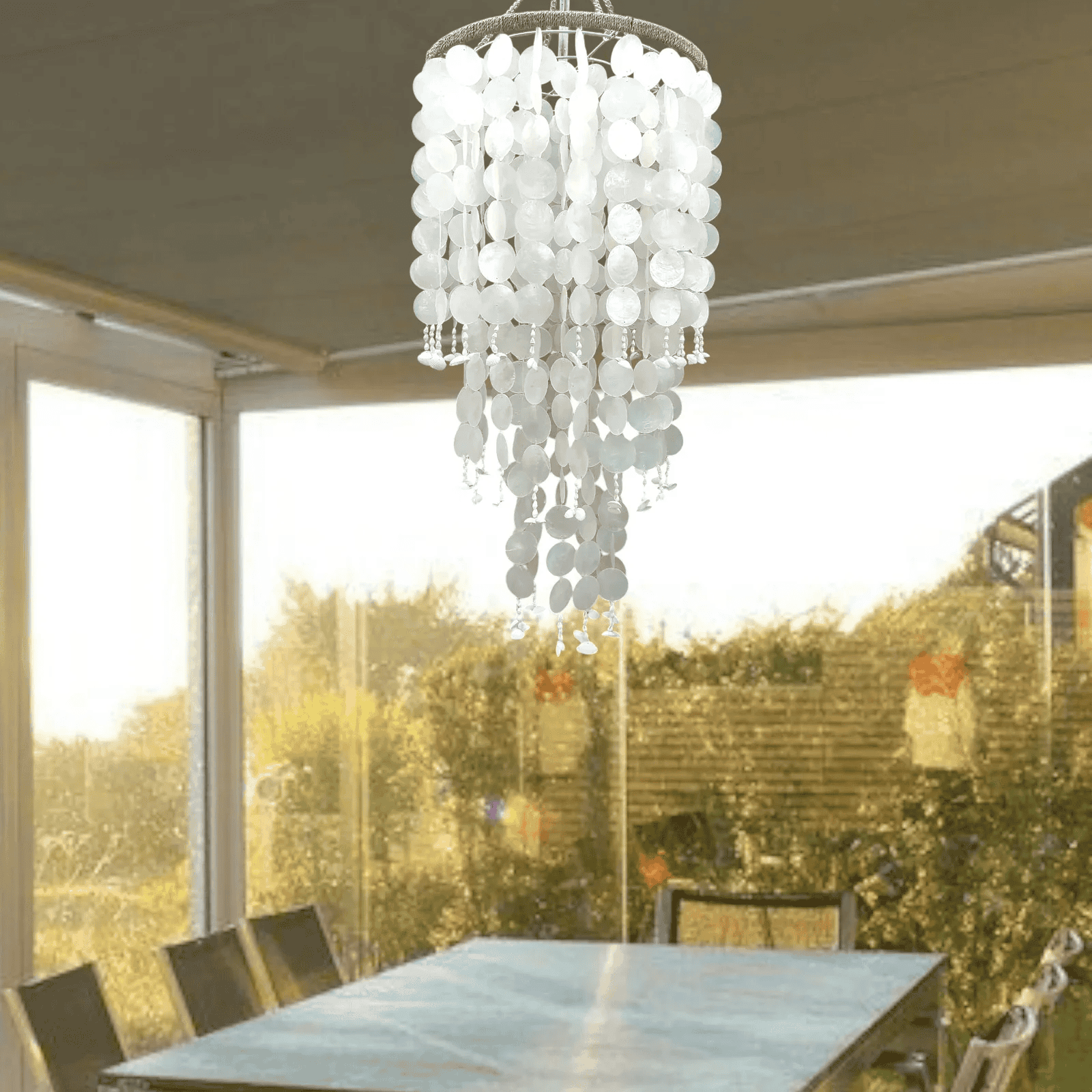 White capiz shell chandelier hanging above a dining table with a view of greenery outside.