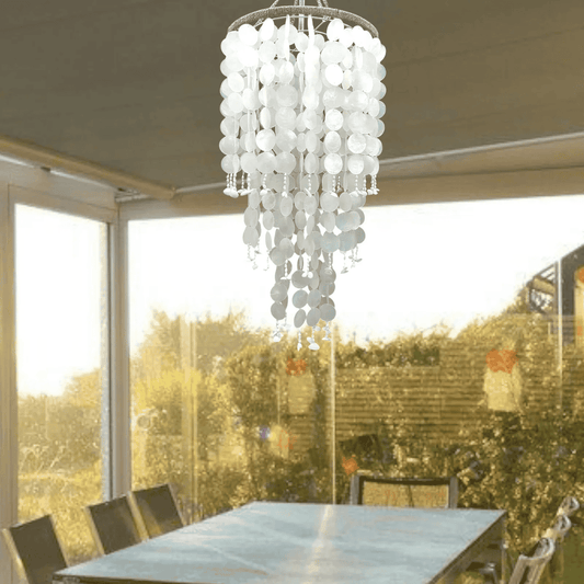 White capiz shell chandelier hanging above a dining table with a view of greenery outside.