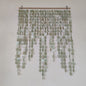 A hanging wall decor piece made of many strands of oval shell-like discs in pale sage green and off-white, suspended from a natural wood rod with two metal clips against a plain light wall.