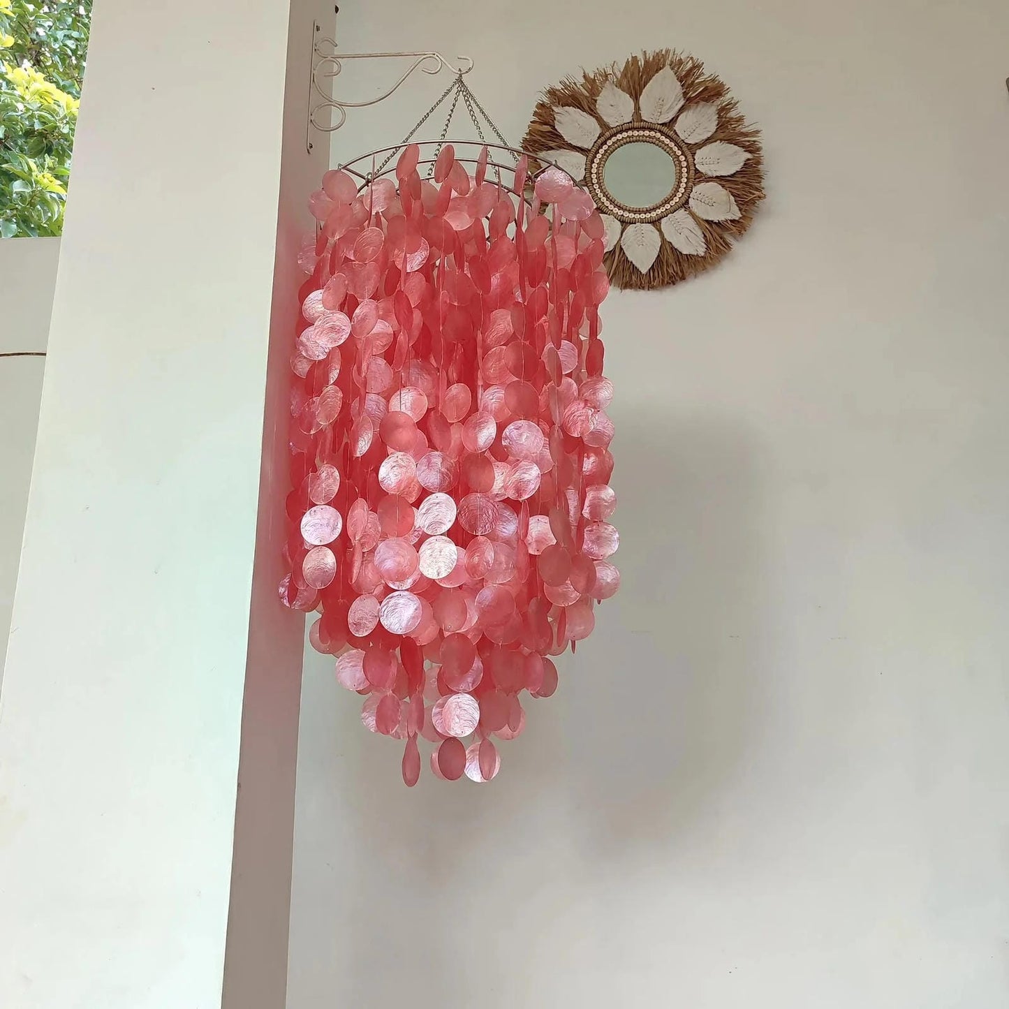 Pink capiz shell chandelier hanging on a wall with a mirror and plant in the background