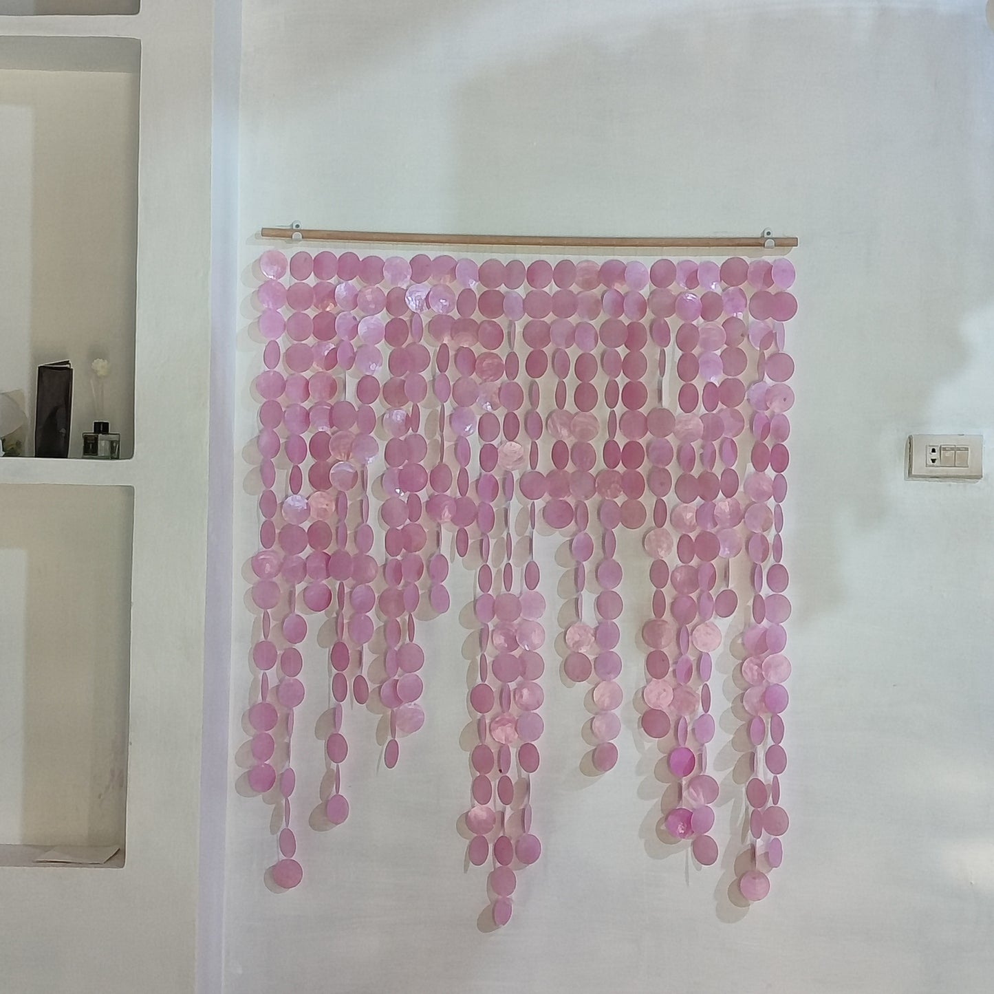 A pink disc bead curtain hanging from a wooden rod on a white wall, with decorative shelving visible on the left.