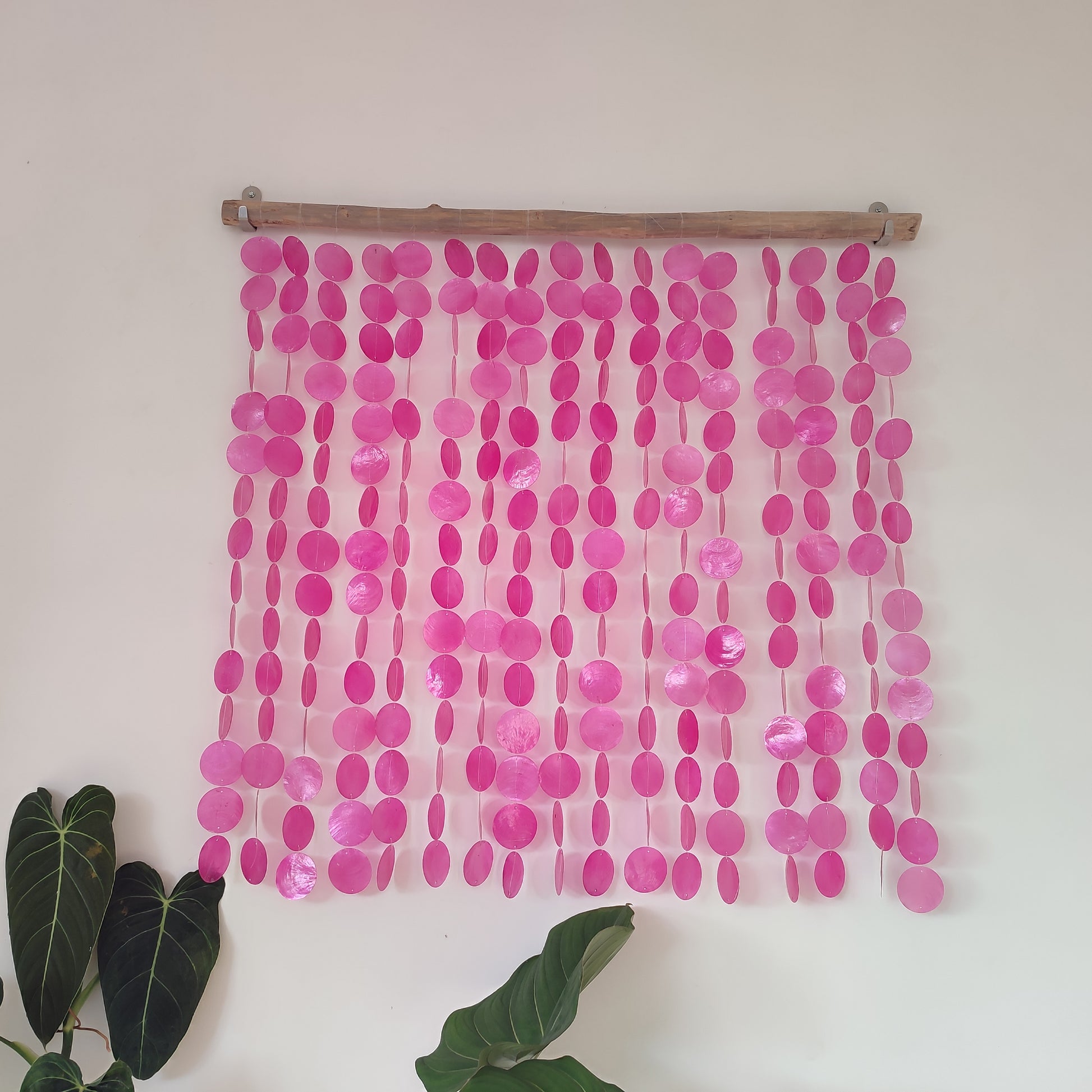 Pink capiznwall hanging on a wooden rod against a white wall with green leaves at the bottom.