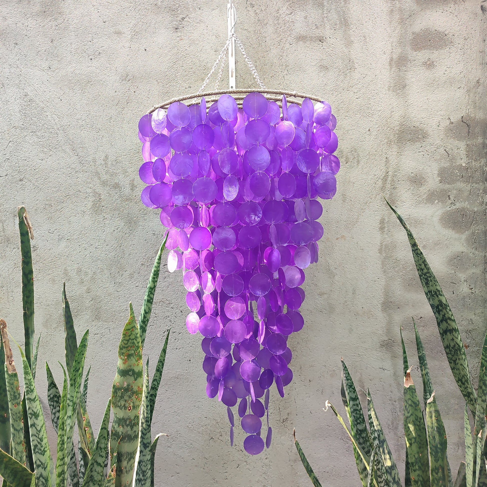 Purple hanging decorative item against a textured wall with plants at the bottom.