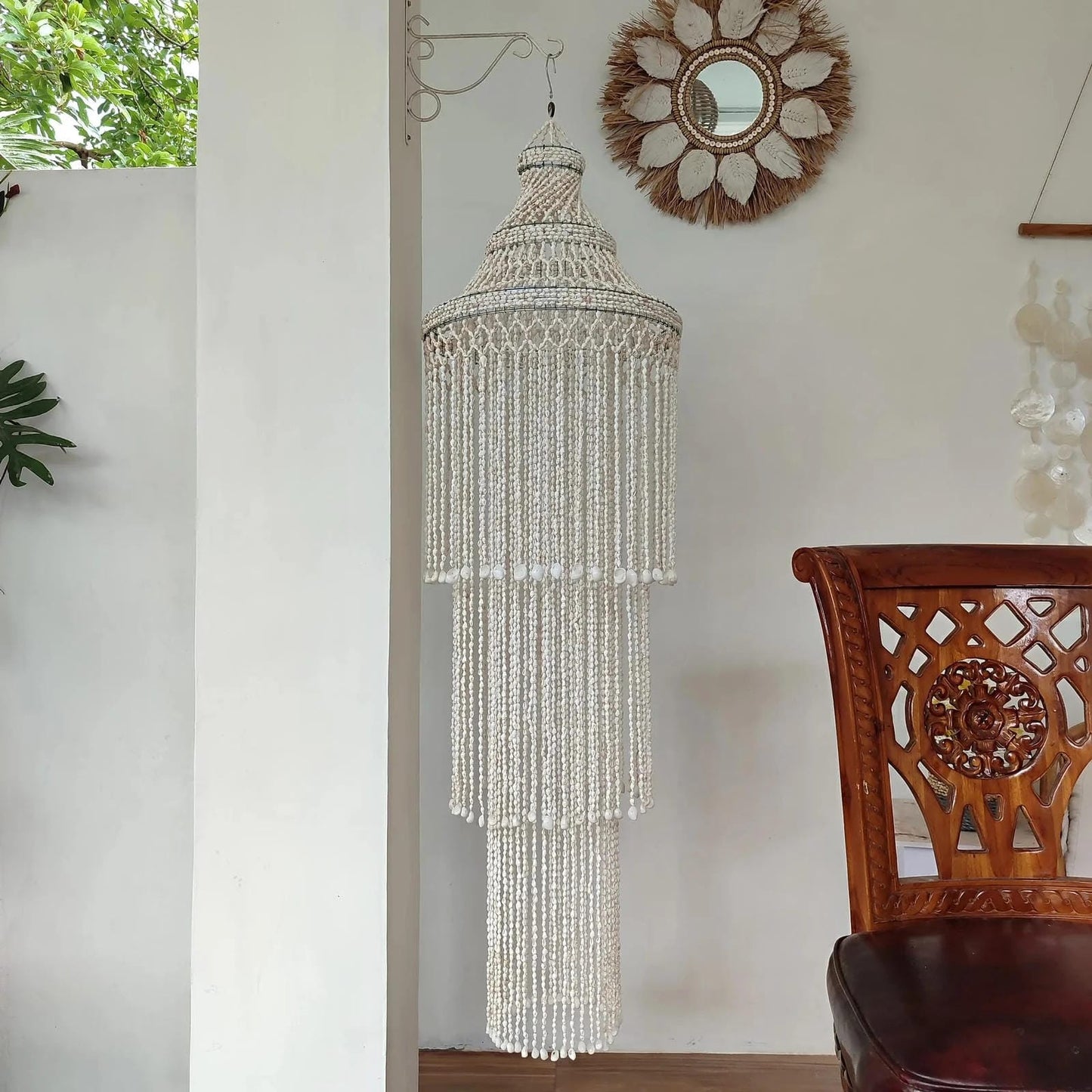 Handmade seashell chandelier hanging in a room with a wooden chair and decorative mirror.