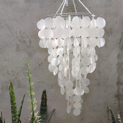 White shell wind chime hanging against a textured wall with plants in the foreground.
