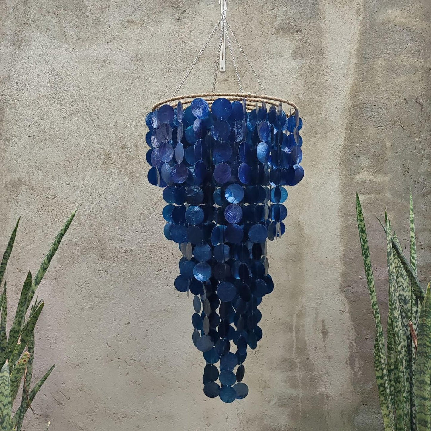 Blue capiz shell chandelier hanging against a textured wall with plants on either side.