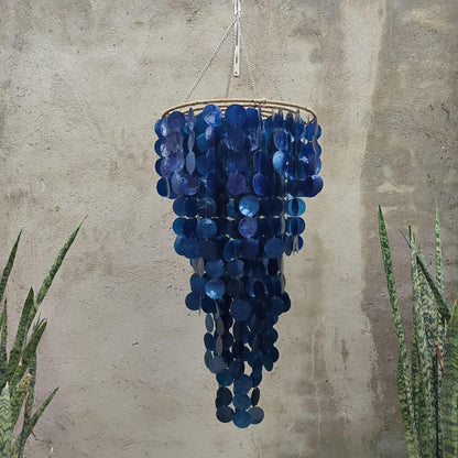 Blue capiz shell chandelier hanging against a textured wall with plants on either side.