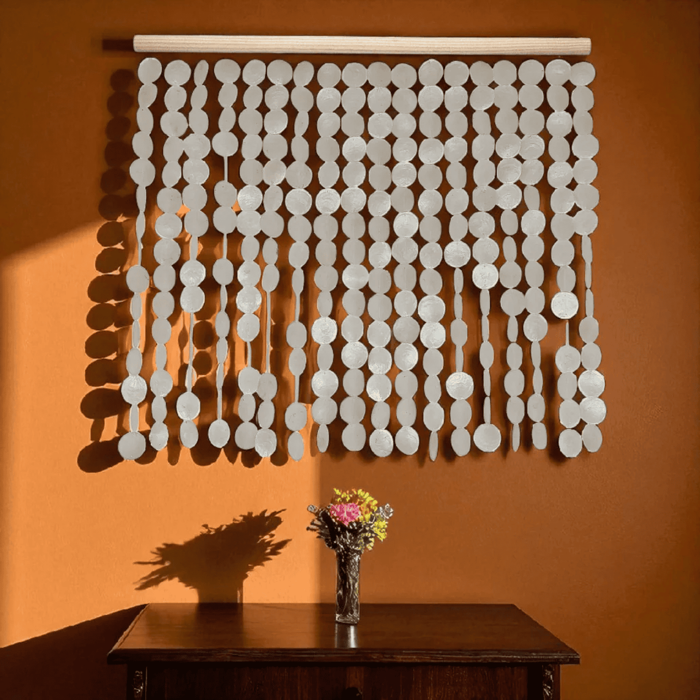 Decorative capiz shell wall hanging on an orange wall, with a vase of flowers on a table below.