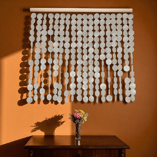 Decorative capiz shell wall hanging on an orange wall, with a vase of flowers on a table below.